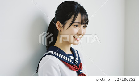 セーラー服を着た、壁際に立つポニーテールで笑顔の女子高生 127186852