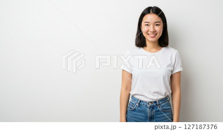 Smiling Asian Woman with Straight Shoulder-Length Hair in White T-Shirt and Blue Jeans Standing Against White Background 127187376