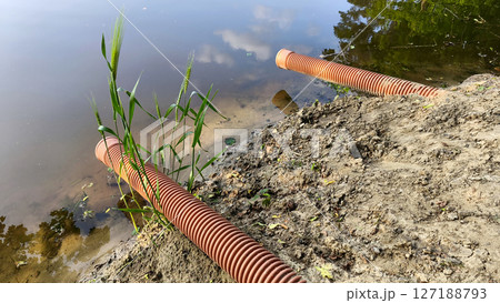 Ribbed drainage pipes descend into a tranquil pond from a worn, sandy shoreline with minimal plant life. Pollution or wastewater outflow . 127188793