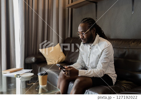 Young African American man relaxing on soft at home, texting messaging on smartphone. man using mobile phone chatting online message, shopping online from home 127192022