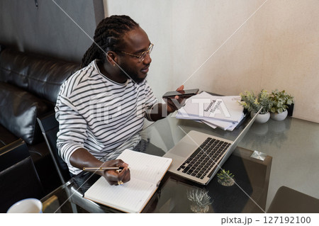 Young African American business man working with laptop and document on desk and talking on mobile at home 127192100