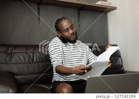 African American business man working with document and laptop while talking on mobile at home on couch. work from home concept 127192109