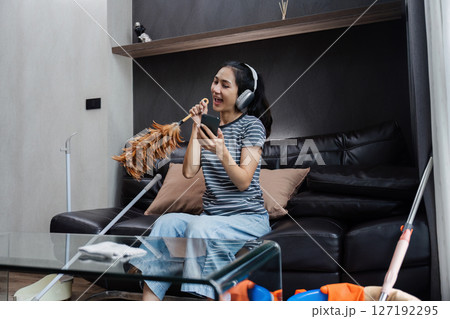 Funny woman Asian teenage cleaning house, doing housework, cleaning the floor with duster and listening to music in wireless headphones 127192295