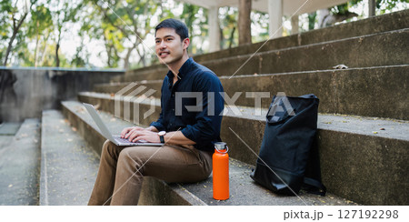 Young businessman working online with laptop while sitting on bench outside of office building 127192298