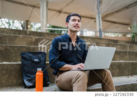 Young businessman working online with laptop while sitting on bench outside of office building 127192330