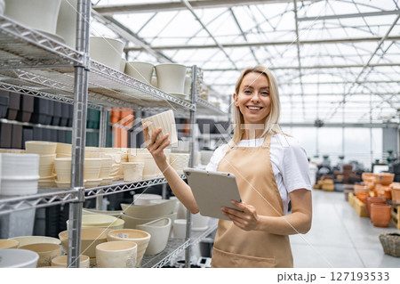 A Caucasian woman working at a plant shop, smiling, holding a flowerpot and a tablet. 127193533
