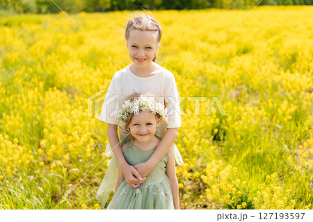 Loving preteen and preschooler sisters in flower crown standing hugging tightly in vibrant field of yellow flowers, enjoying sunny summer day together, looking at camera with happy expression. Loving preteen and preschooler sisters in flower crown standing hugging tightly in vibrant field of yellow flowers, enjoying sunny summer day together, looking at camera with happy expression. 127193597