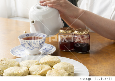 焼きたての紅茶のスコーンにジャムを乗せて手で持ち上げる　紅茶のスコーンとティーポットとティーカップ 127193711