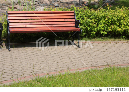 Wooden bench, wrought iron trash can near tile path in park. Green lawn, bushes and trees in summer on sunny day. Background for design. Ecology and nature. Landscape design.  127195111