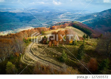 Aerial view of scenic landscape in autumn, showcasing rolling hills adorned with vibrant fall foliage. Winding dirt path weaves through trees, leading towards distant valleys and villages. 127203572