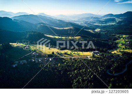 Aerial view of densely forested mountains stretching into distance under clear blue sky with scattered clouds. Landscape reveals valleys and distant fields, showcasing natural beauty of region. 127203605
