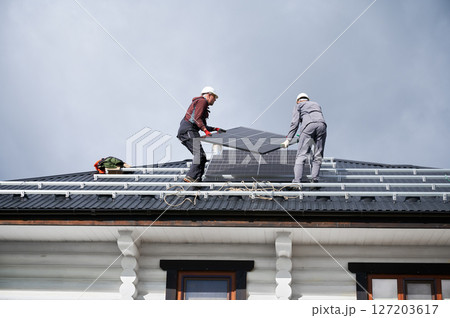 Builders installing solar panel system on roof of house. Men workers in helmets carrying photovoltaic solar module outdoors. Concept of alternative and renewable energy. Builders installing solar panel system on roof of house. Men workers in helmets carrying photovoltaic solar module outdoors. Concept of alternative and renewable energy. 127203617