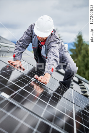 Man roofer mounting photovoltaic solar panels on roof of house. Engineer in helmet installing solar module system with help of hex key. Concept of alternative, renewable energy. Man roofer mounting photovoltaic solar panels on roof of house. Engineer in helmet installing solar module system with help of hex key. Concept of alternative, renewable energy. 127203618
