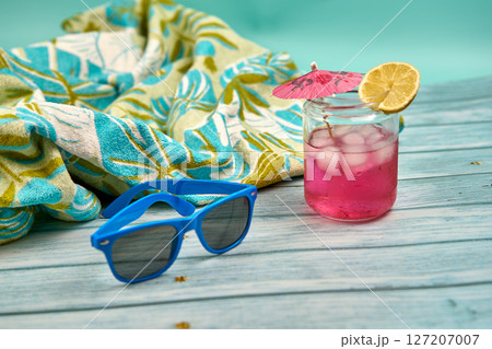 Refreshing summer drink with lots of ice an umbrella and a slice of lemon, blue sunglasses and tropical patterned beach towel on a wooden table and a greenish background. 127207007
