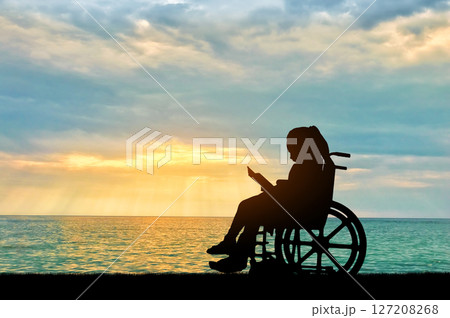 Silhouette of a disabled child girl sitting in a wheelchair reading a book on a sea sunset background 127208268