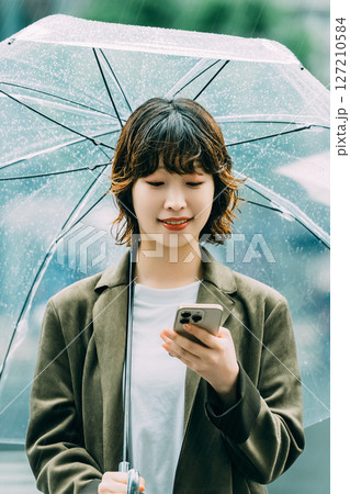 雨の中、傘をさしてスマホを使う女性（梅雨・6月） 127210584