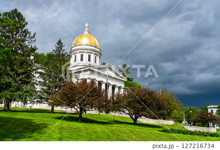 Front view of the Vermont State House in Montpelier, featuring a gold dome and neoclassical architecture, surrounded by lush trees on a bright spring day 127216734