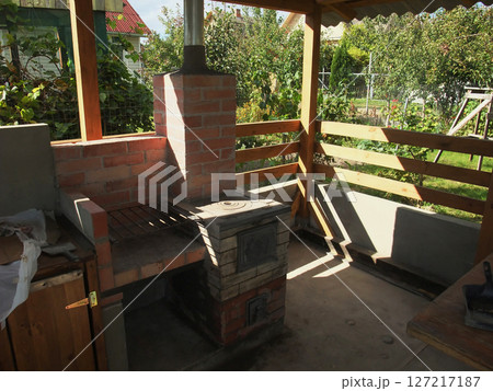 A cozy outdoor cooking area features a brick stove and wooden seating. Sunlight filters through a shelter, highlighting the rustic charm. The garden setting offers greenery and tranquility. 127217187