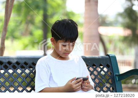 Young boy engrossed in smartphone while sitting on park bench, showcasing technology's impact on childhood and leisure activities in a natural environment 127218020