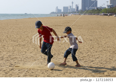 Two Young Boys Playing Soccer on Sandy Beach with Sunny Sky and Cityscape in the Background, Capturing Joy of Childhood and Outdoor Activity in a Coastal Setting 127218053