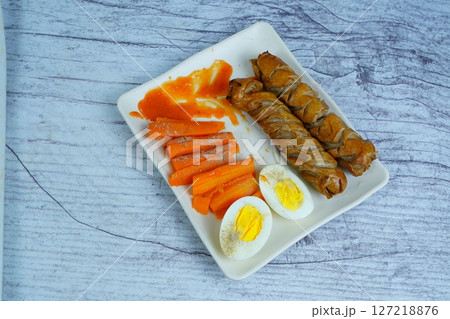 A healthy and delicious meal of sausages, boiled eggs, carrots, and chili sauce on a white plate. 127218876