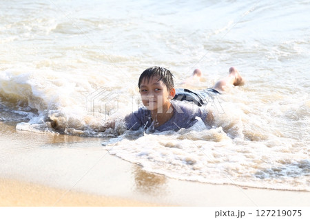 Young boy enjoying a sunny day at the beach, playing in the gentle ocean waves while lying on his stomach on the warm sandy shore, filled with joy and excitement. 127219075