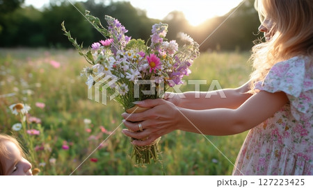 Child presents a bouquet of wildflowers in a sunlit meadow during golden hour near a family member 127223425