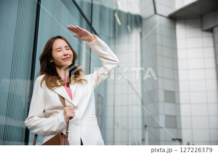 Happy woman wearing white suit jacket with brown bag at sunlight outdoors. Portrait of confidence young businesswoman standing outside office building in city raise your hand to shade sun Happy woman wearing white suit jacket with brown bag at sunlight outdoors. Portrait of confidence young businesswoman standing outside office building in city raise your hand to shade sun 127226379