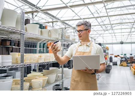 A Caucasian man inspects a flower pot in a shop with multiple flowerpots on shelves, while holding a laptop. A Caucasian man inspects a flower pot in a shop with multiple flowerpots on shelves, while holding a laptop. 127226903