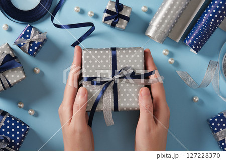 Hands hold a silver-colored gift box. Around it are gifts and ribbons in blue and silver colors. Hands hold a silver-colored gift box. Around it are gifts and ribbons in blue and silver colors. 127228370