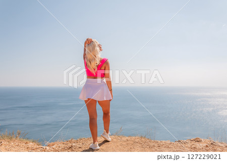 A blonde woman stands on a rocky beach, looking out at the ocean 127229021