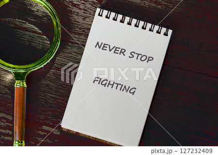 The concept of inspirational and motivational. NEVER STOP FIGHTING written on a notebook on a dark background. There's a magnifying glass next to it 127232409