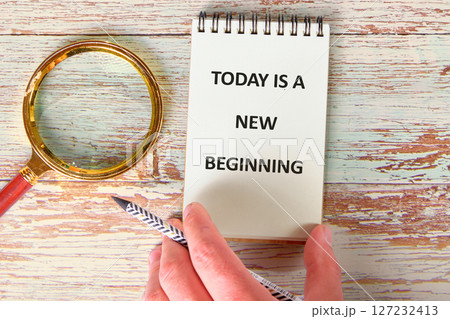 Today is a New Beginning text message written on a notebook. There is a magnifying glass on the wooden surface nearby 127232413