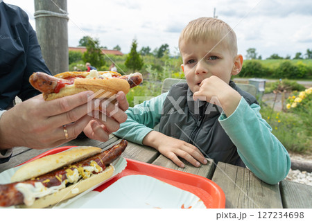 Young boy sitting at outdoor table looks reluctant as adult offers him a large hot dog with condiments. The setting is a picnic area with natural surroundings. Young boy sitting at outdoor table looks reluctant as adult offers him a large hot dog with condiments. The setting is a picnic area with natural surroundings. 127234698