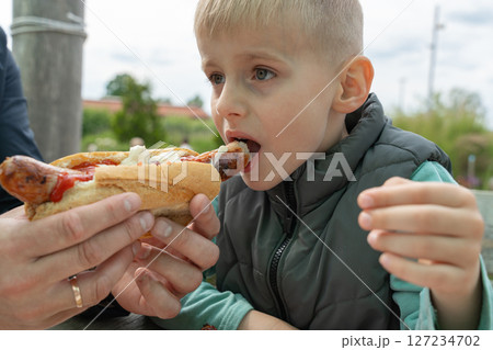 Young child eagerly biting into a hot dog while seated outdoors. The child shows a focused expression as they enjoy their meal, creating a moment of satisfaction and enjoyment. 127234702