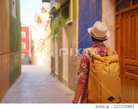 Woman in dress strolls through colorful streets of Spanish coastal town of La Vila Joiosa . sunny winter atmosphere highlights charm of Mediterranean architecture and quiet seaside life, back view 127235661
