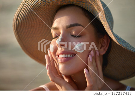 Portrait of beautiful smiling woman applying sunscreen on her face while looking at camera at the beach. Female smears sunscreen lotion on skin. Skin Protection and dermatology Portrait of beautiful smiling woman applying sunscreen on her face while looking at camera at the beach. Female smears sunscreen lotion on skin. Skin Protection and dermatology 127237413