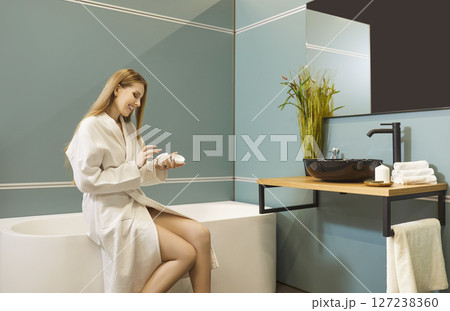 Smiling young woman in bathrobe sitting on bathtub and applying skincare cream after morning shower. Smiling young woman in bathrobe sitting on bathtub and applying skincare cream after morning shower. 127238360