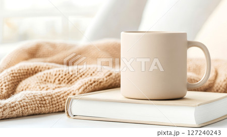 Beige ceramic mug standing on book with knitted blanket on white table in cozy bright bedroom Beige ceramic mug standing on book with knitted blanket on white table in cozy bright bedroom 127240243