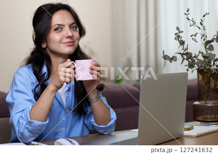 Beautiful woman in blue shirt drinking coffee working on laptop computer while sitting at home office 127241632