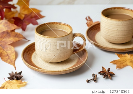 Spicy cappuccino two cups. Spice latte with art foam, cinnamon stick and anise stars. Autumn leaves. Warm Coffee on a white. Cozy fall drink on a white table. Inviting atmosphere. Mug of warming drink 127248562