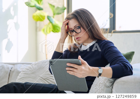 Serious middle aged woman looking at digital tablet sitting on sofa at home 127249859