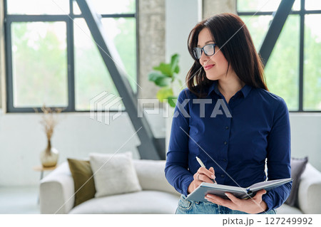 Portrait of smiling business woman with notebook in office with couch Portrait of smiling business woman with notebook in office with couch 127249992