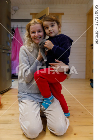 Heartwarming Moment of Joy: A Woman Smiling Happily While Embracing a Young Girl Holding a Cute Gray Kitten in Cozy Indoor Setting with Natural Wood Elements Heartwarming Moment of Joy: A Woman Smiling Happily While Embracing a Young Girl Holding a Cute Gray Kitten in Cozy Indoor Setting with Natural Wood Elements 127250095