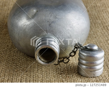 Old soldier's aluminum canteen with lid on a chain on burlap. Thermos for water. Empty canteen with an unscrewed stopper. Old soldier's aluminum canteen with lid on a chain on burlap. Thermos for water. Empty canteen with an unscrewed stopper. 127251489
