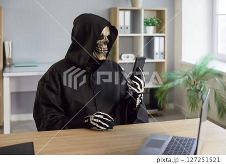 Man in death costume with black hood and skeleton mask sitting in office looking at mobile phone Man in death costume with black hood and skeleton mask sitting in office looking at mobile phone 127251521