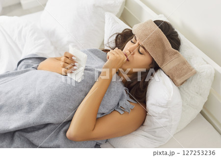 Sick young woman with wet towel on head lying in bed, suffering from cold or flu virus and coughing. Sick young woman with wet towel on head lying in bed, suffering from cold or flu virus and coughing. 127253236