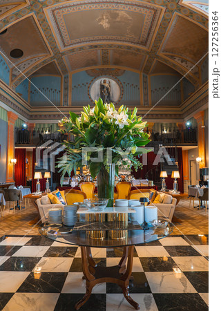 Opulent lounge with high decorated ceiling, central mural, floral centerpiece, plush seating, checkered marble floor, red curtains, and chandeliers. Opulent lounge with high decorated ceiling, central mural, floral centerpiece, plush seating, checkered marble floor, red curtains, and chandeliers. 127256364