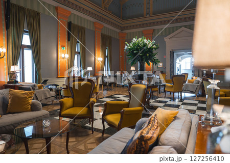 Spacious lounge with high ceilings, tall windows, green and gold drapes, orange columns, checkered marble floor, plush seating, and floral centerpiece. 127256410