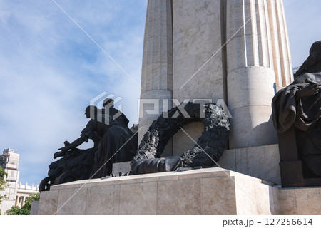 A tall stone column with intricate sculptures at its base, including human figures and a wreath, set against a bright blue sky in Budapest, Hungary. 127256614